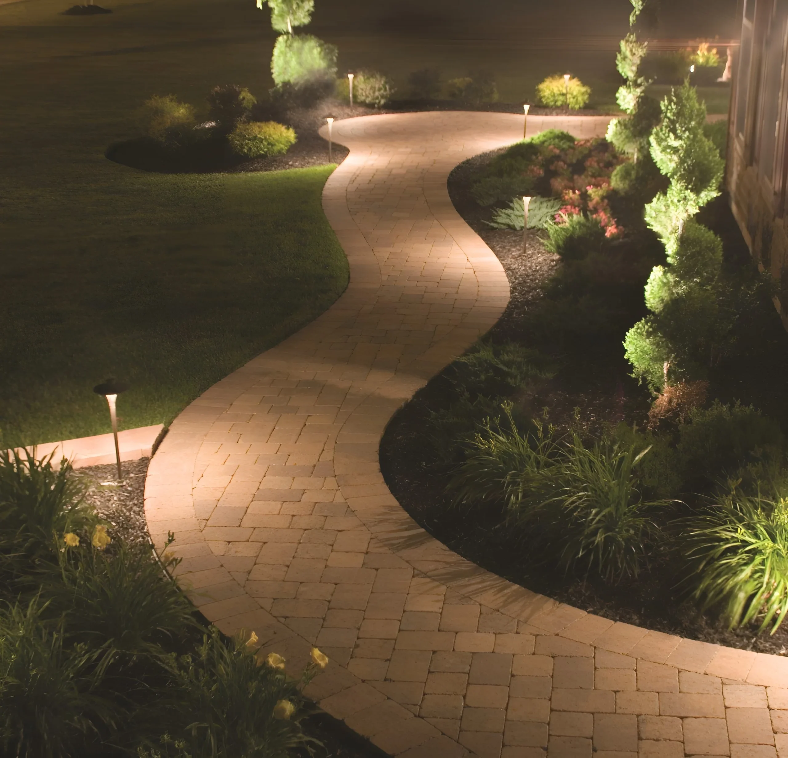 Path lights line a winding paver walk through planted beds, landscape lighting for homes in Scarsdale.