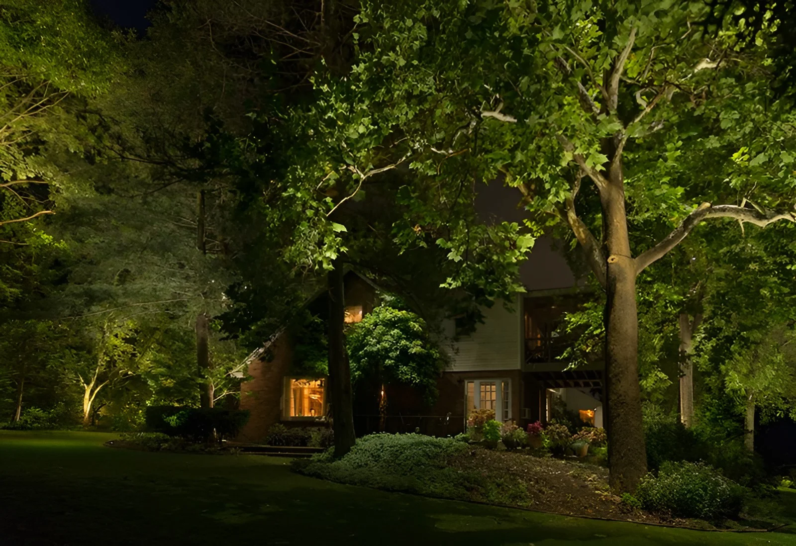 Large uplit trees shade a brick and clapboard house at night, with a deep lawn in front.
