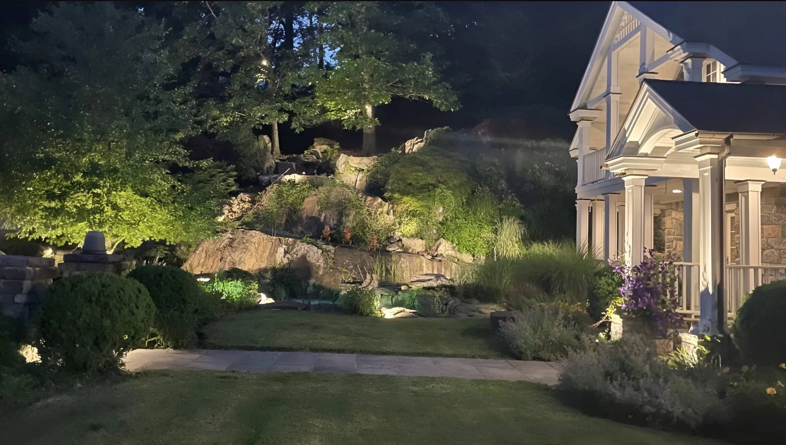 Uplit trees and a rock garden glow beside a columned porch, landscape lighting for homes in Rye.