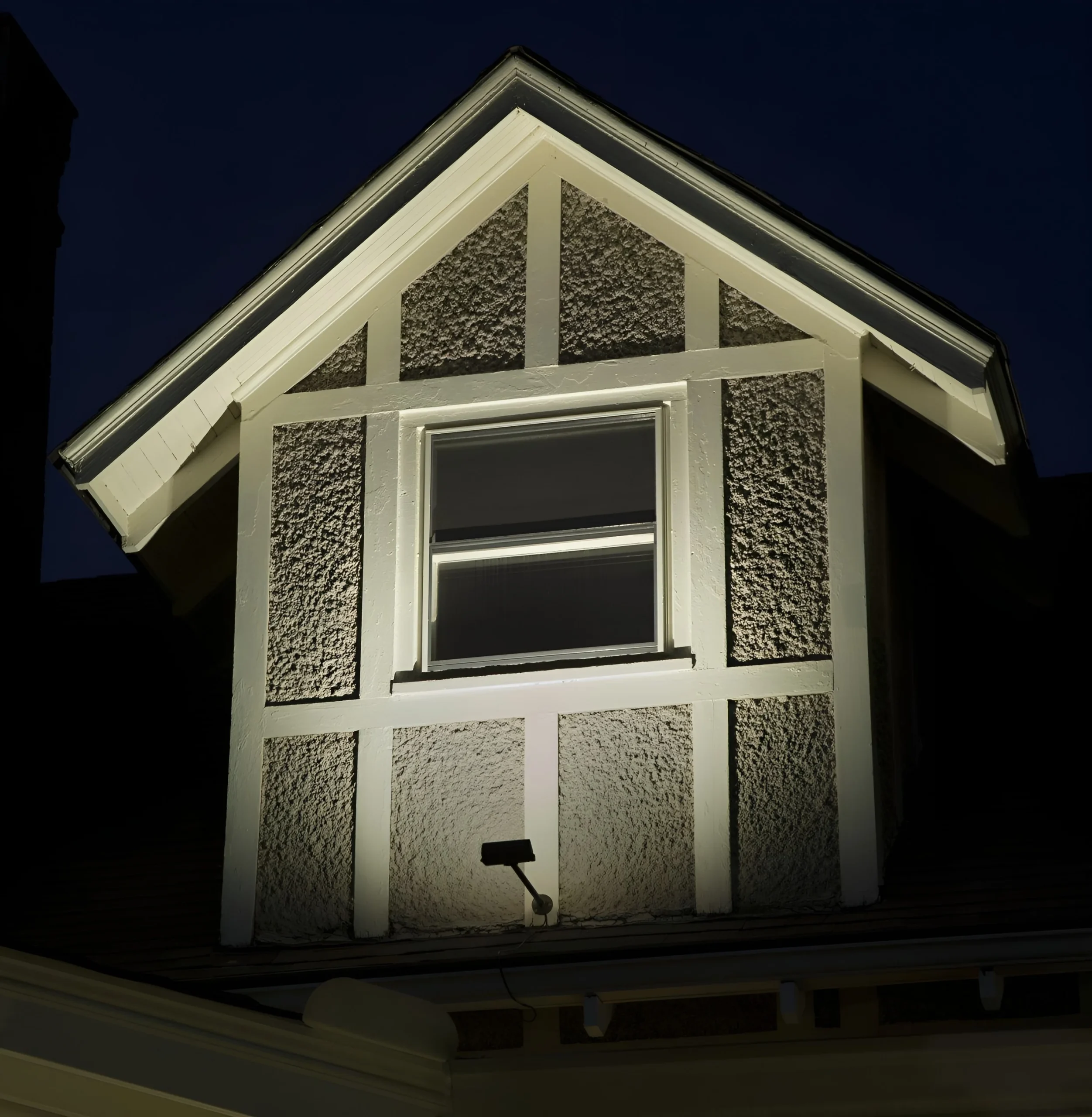 Light from a small fixture grazes the stucco and half-timbering of a Tudor dormer at night.