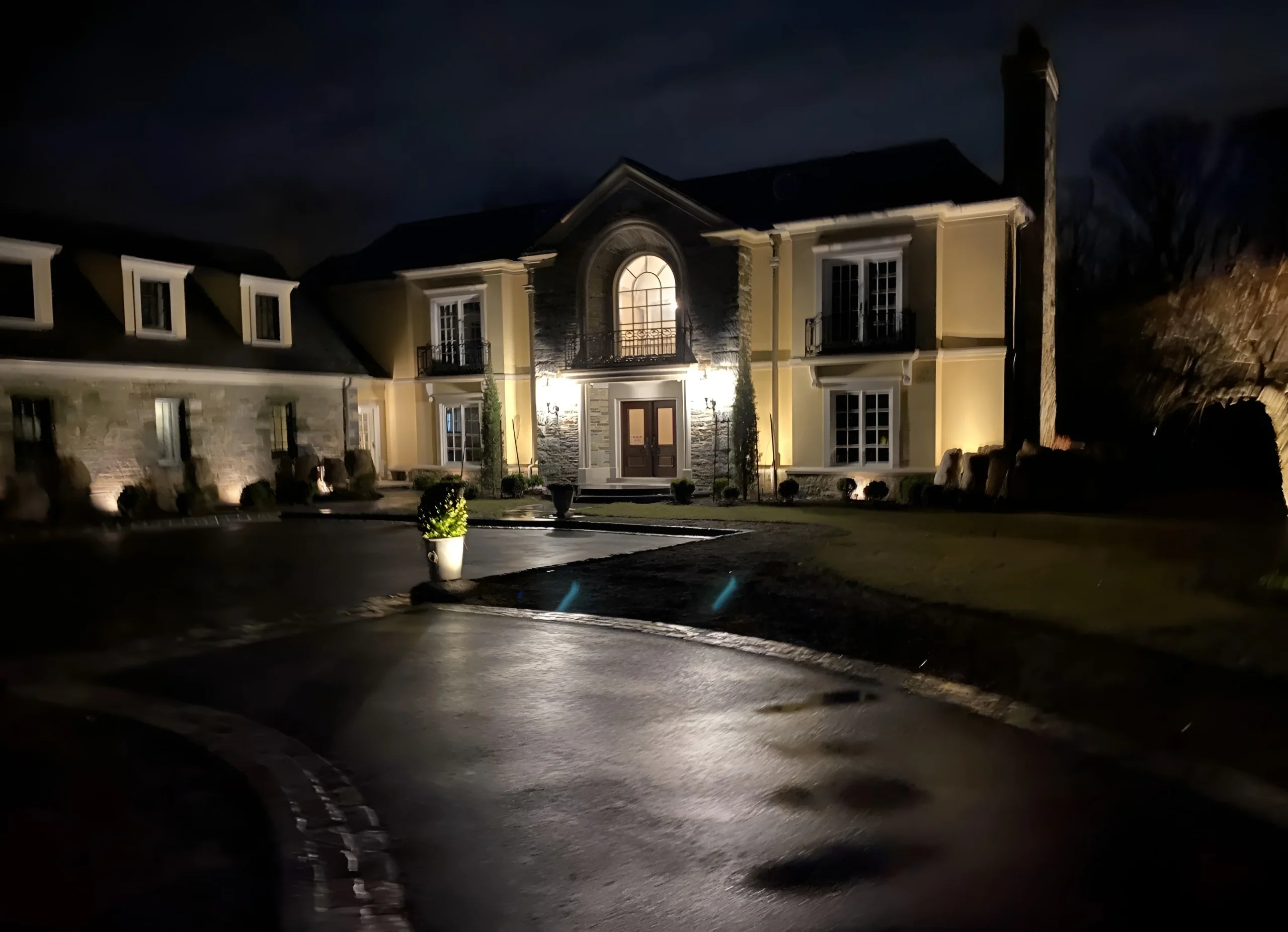 Warm light washes the stone entry and stucco walls of a large house at the end of a wet drive at night.
