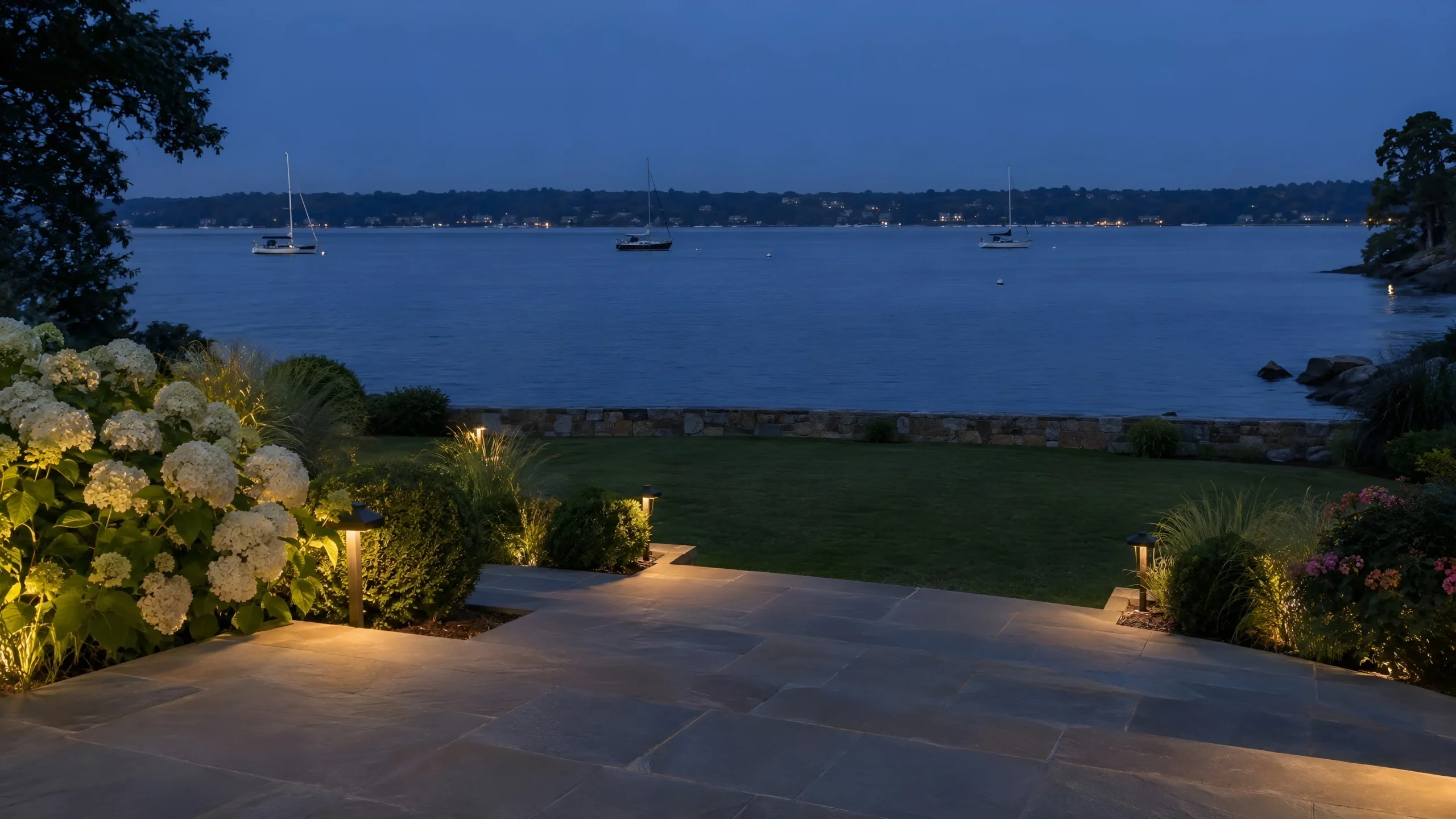 Path lights and uplit hydrangeas line a bluestone terrace above a harbor at dusk, with boats moored beyond.