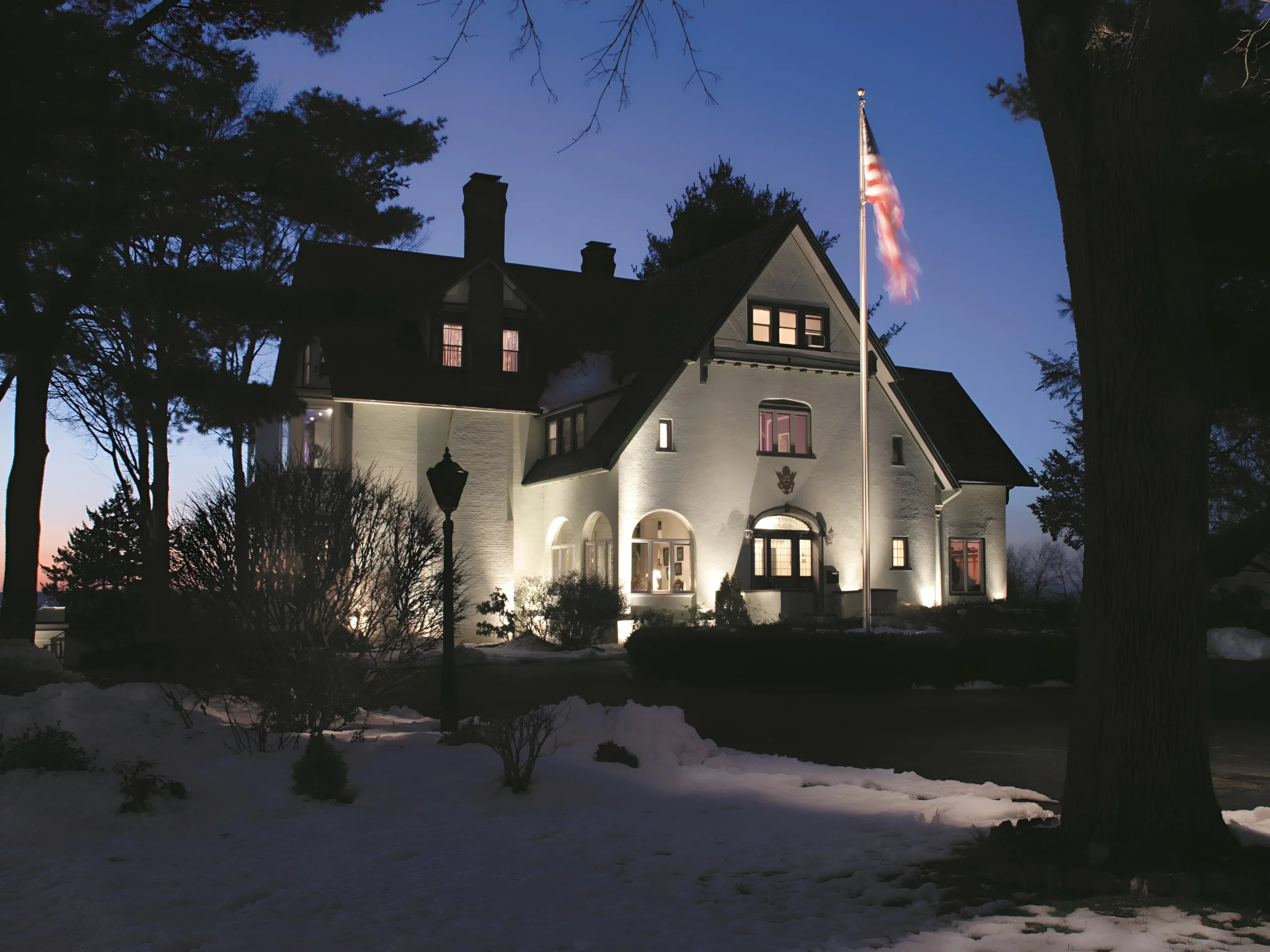 Snow covers the lawn below a white stucco house, its walls and arched windows washed by uplights.