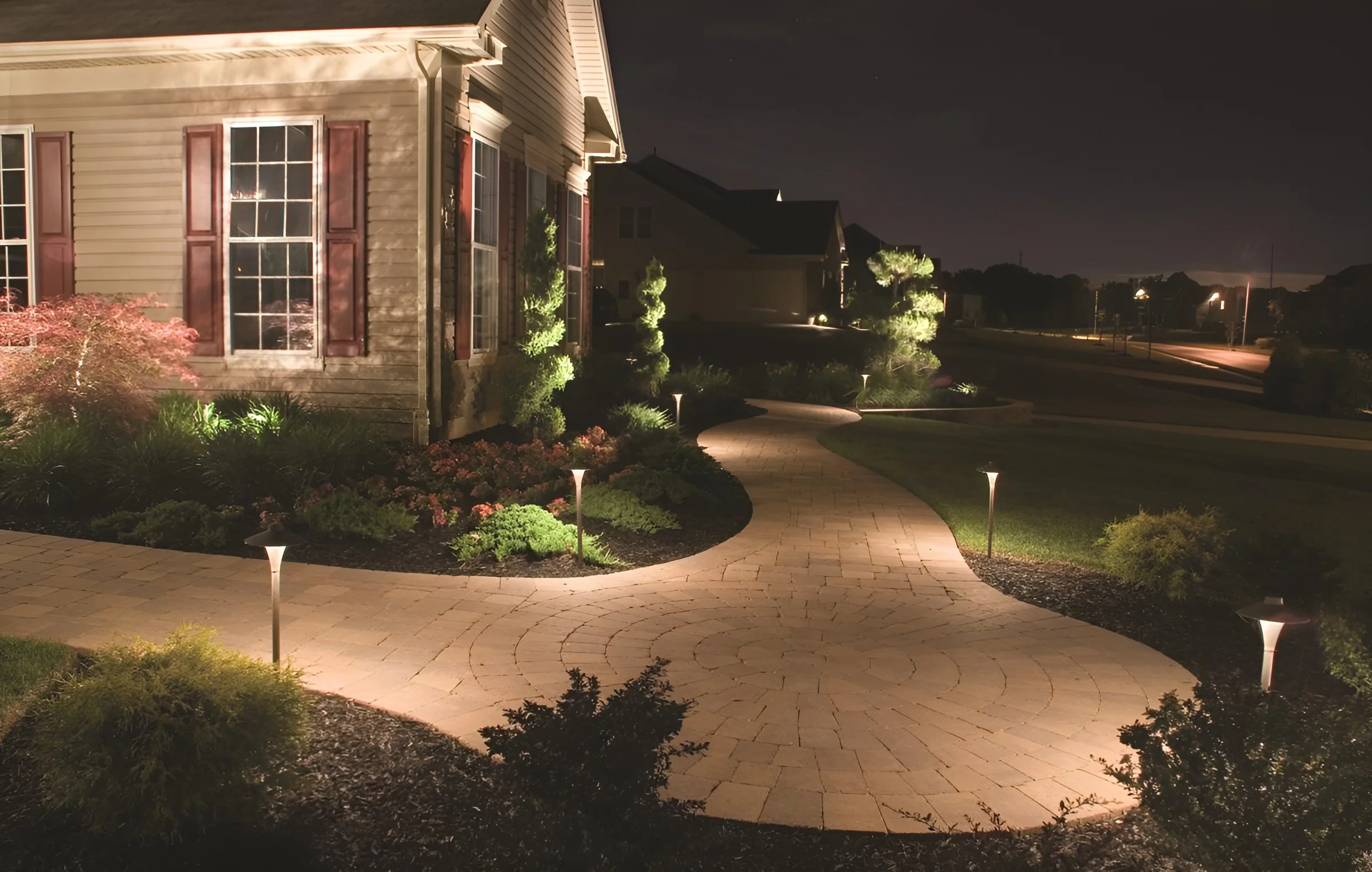 Path lights line a curving paver walk to a sided house at night, with topiaries and shrubs lit in the beds.