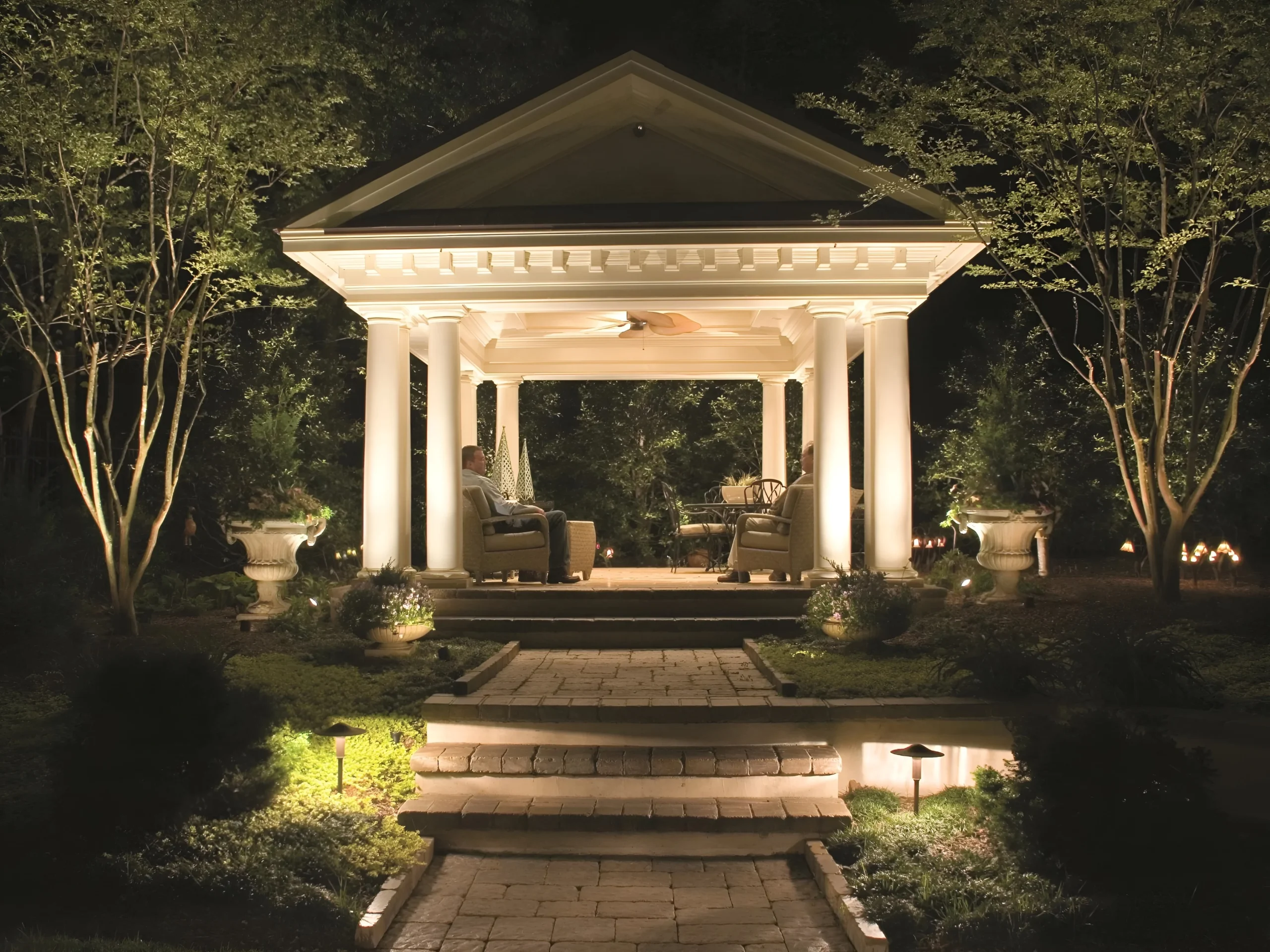A garden pavilion with white columns glows at night among uplit trees, with two people seated inside.